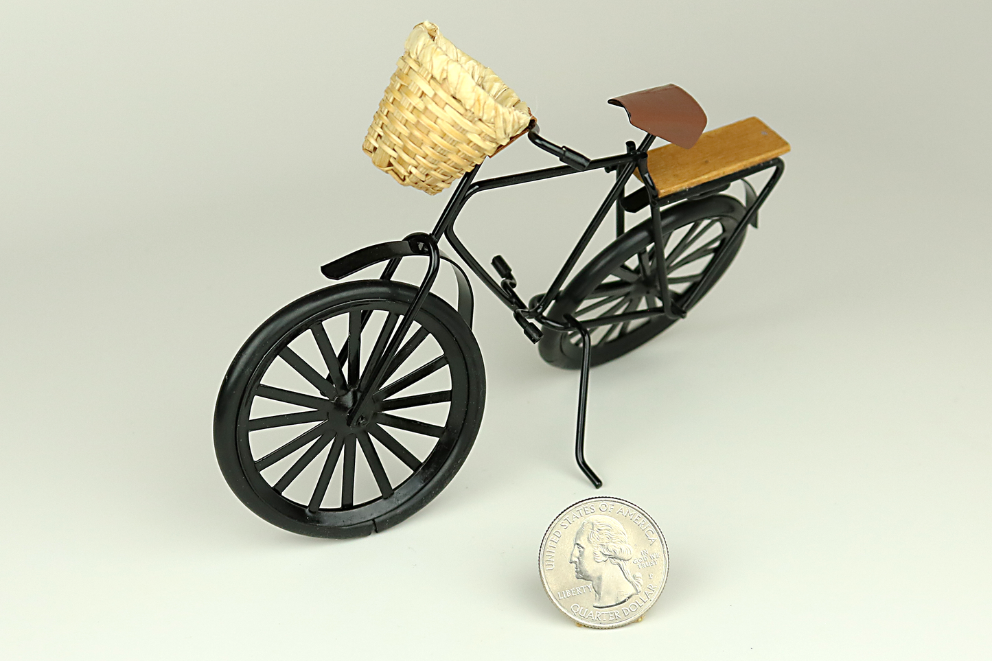 Miniature black bicycle with a basket next to a quarter for scale on a light gray background.