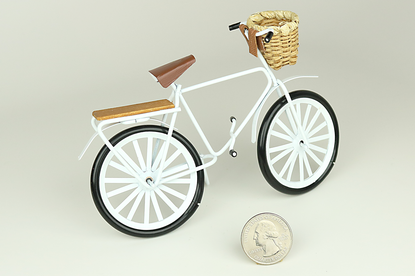 Miniature white bicycle with a basket next to a quarter for scale on a light gray background.