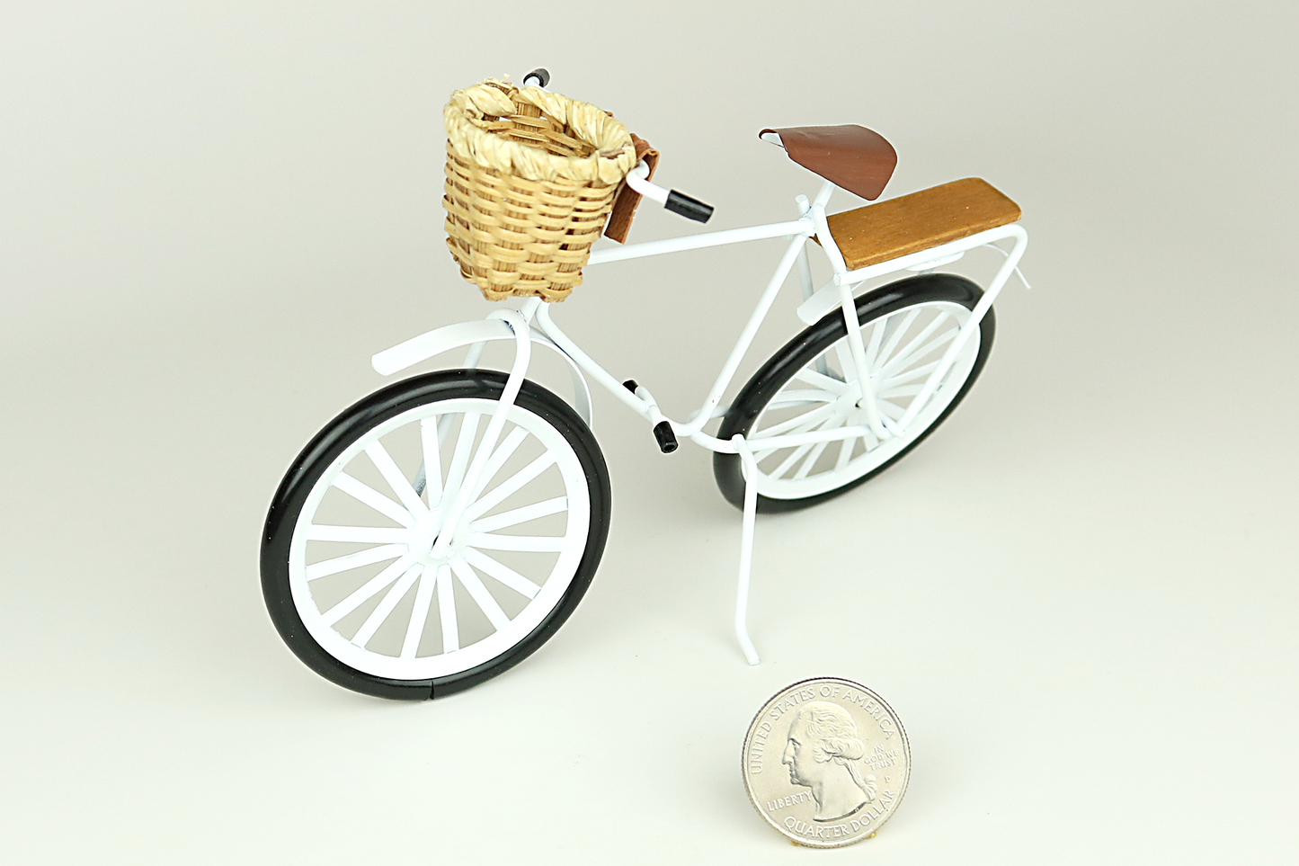 Miniature white bicycle with a basket next to a quarter for scale on a light gray background.