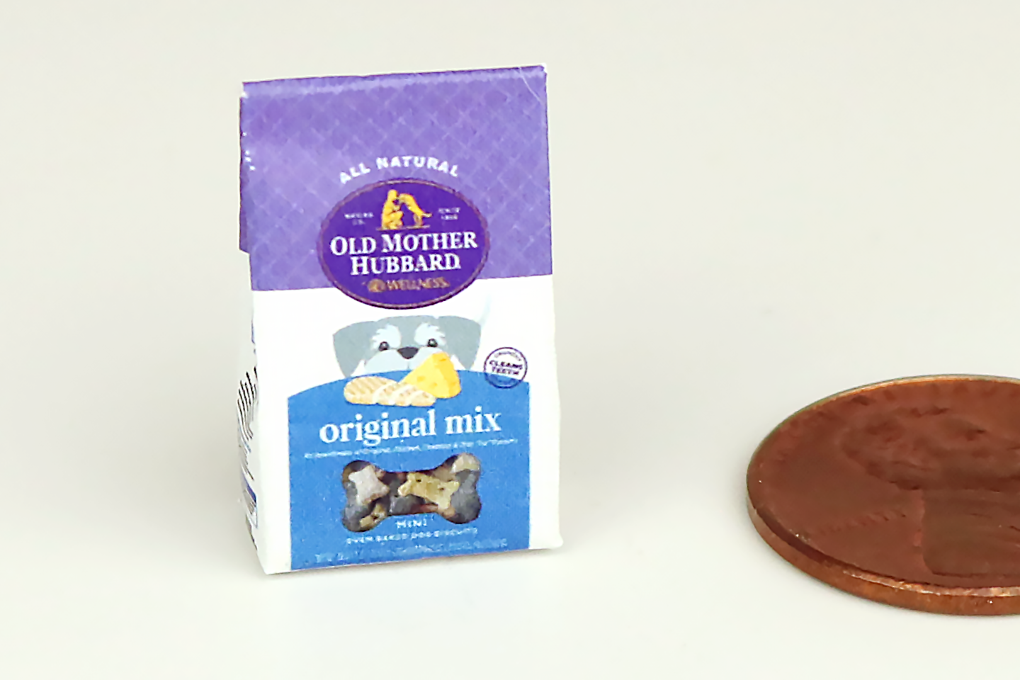 A miniature bag of dog treats with a cartoon dog's face on it, a small "window" showing off the treats, and a purple and blue theme. Pictured next to a penny for scale.