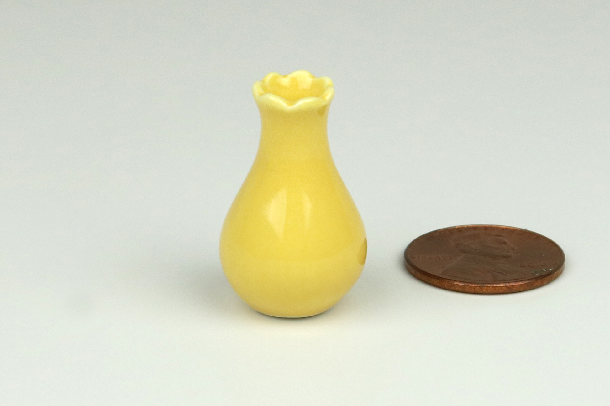 Miniature yellow ceramic vase, wider on the bottom, tapered in at the top, with a scalloped top edge, next to a penny on a white background