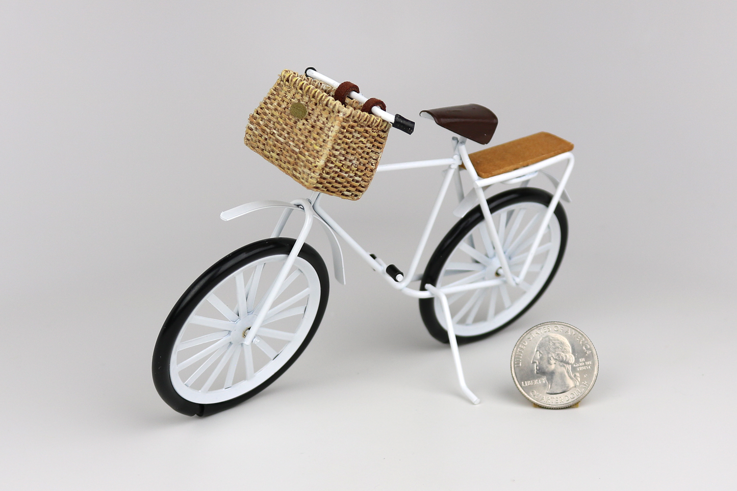 Miniature white bicycle with a basket next to a quarter for scale on a light gray background.