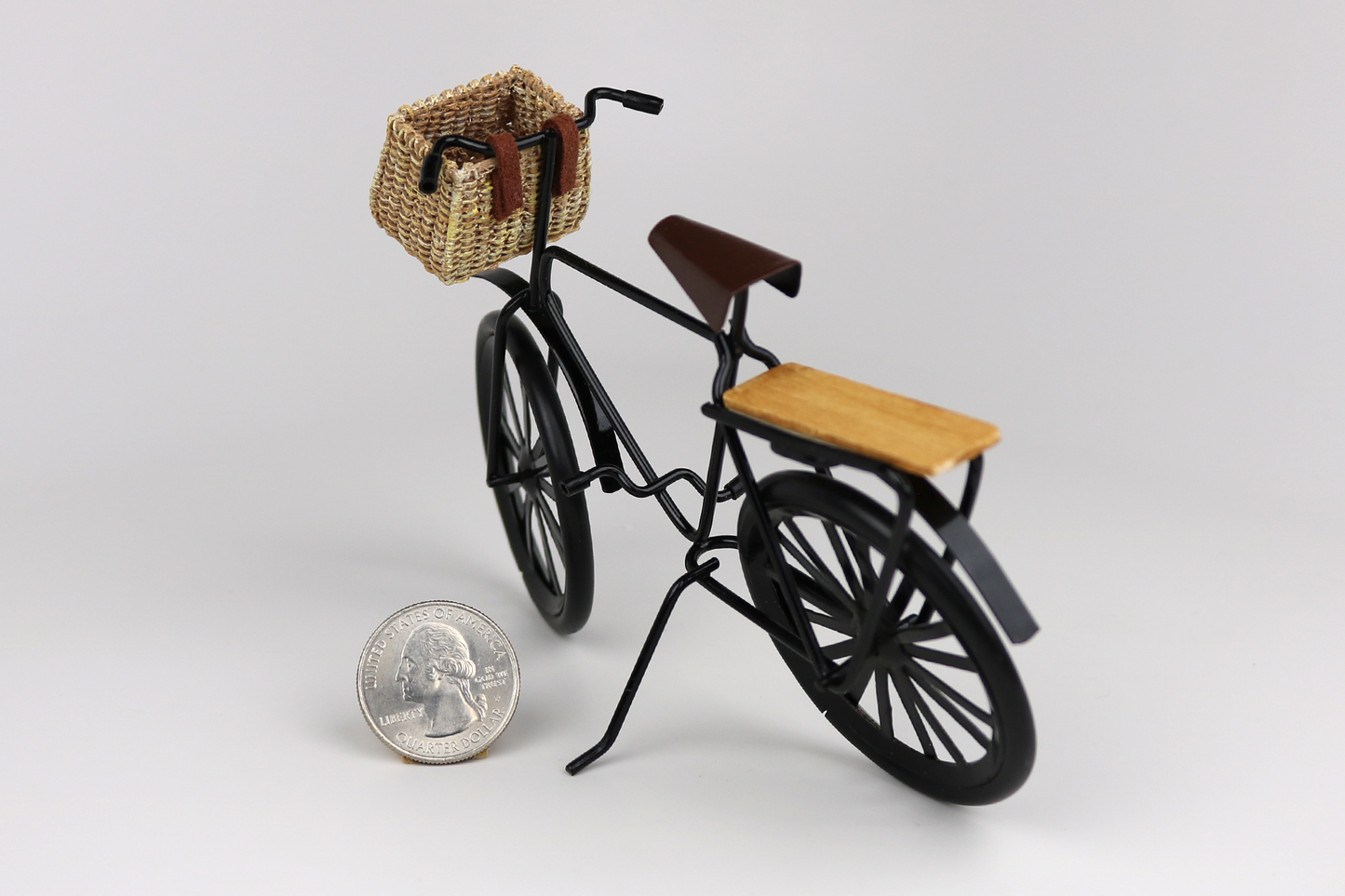 Miniature black bicycle with a basket next to a quarter for scale on a light gray background.