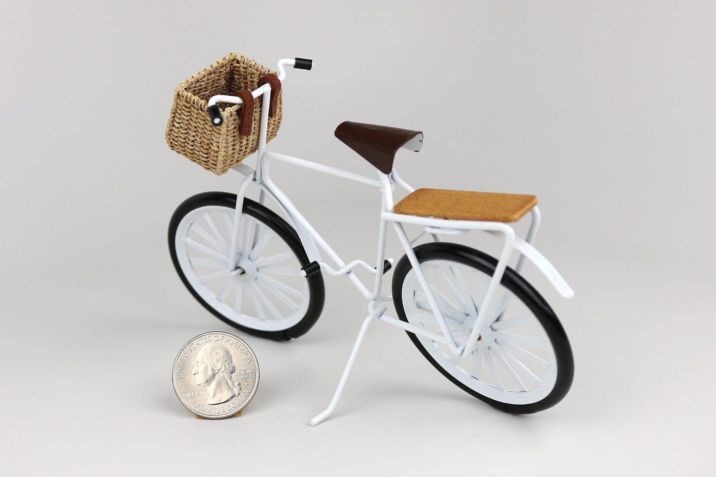 Miniature white bicycle with a basket next to a quarter for scale on a light gray background.