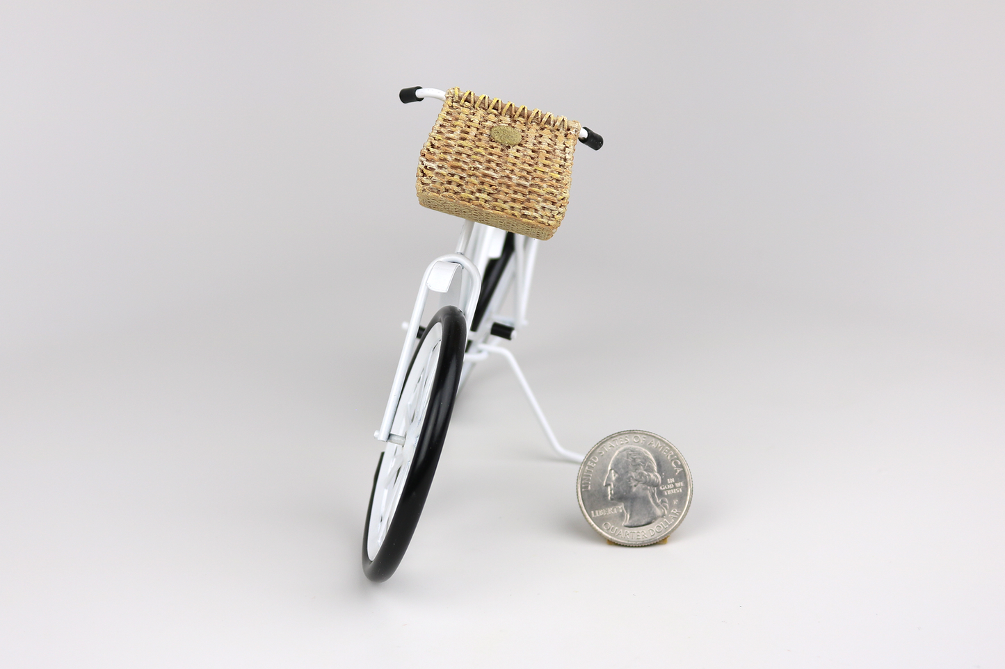 Miniature white bicycle with a basket next to a quarter for scale on a light gray background.