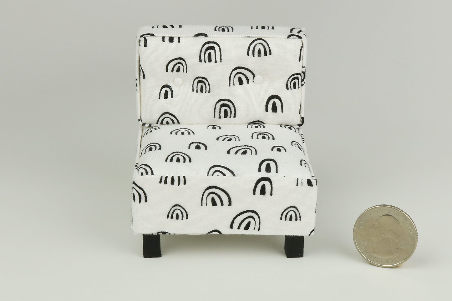 Miniature mid-century modern style side chair with no arms, in a white fabric with black rainbow-shaped patterns throughout, a tufted back with two buttons, and square block walnut legs, next to a quarter for scale on a white background