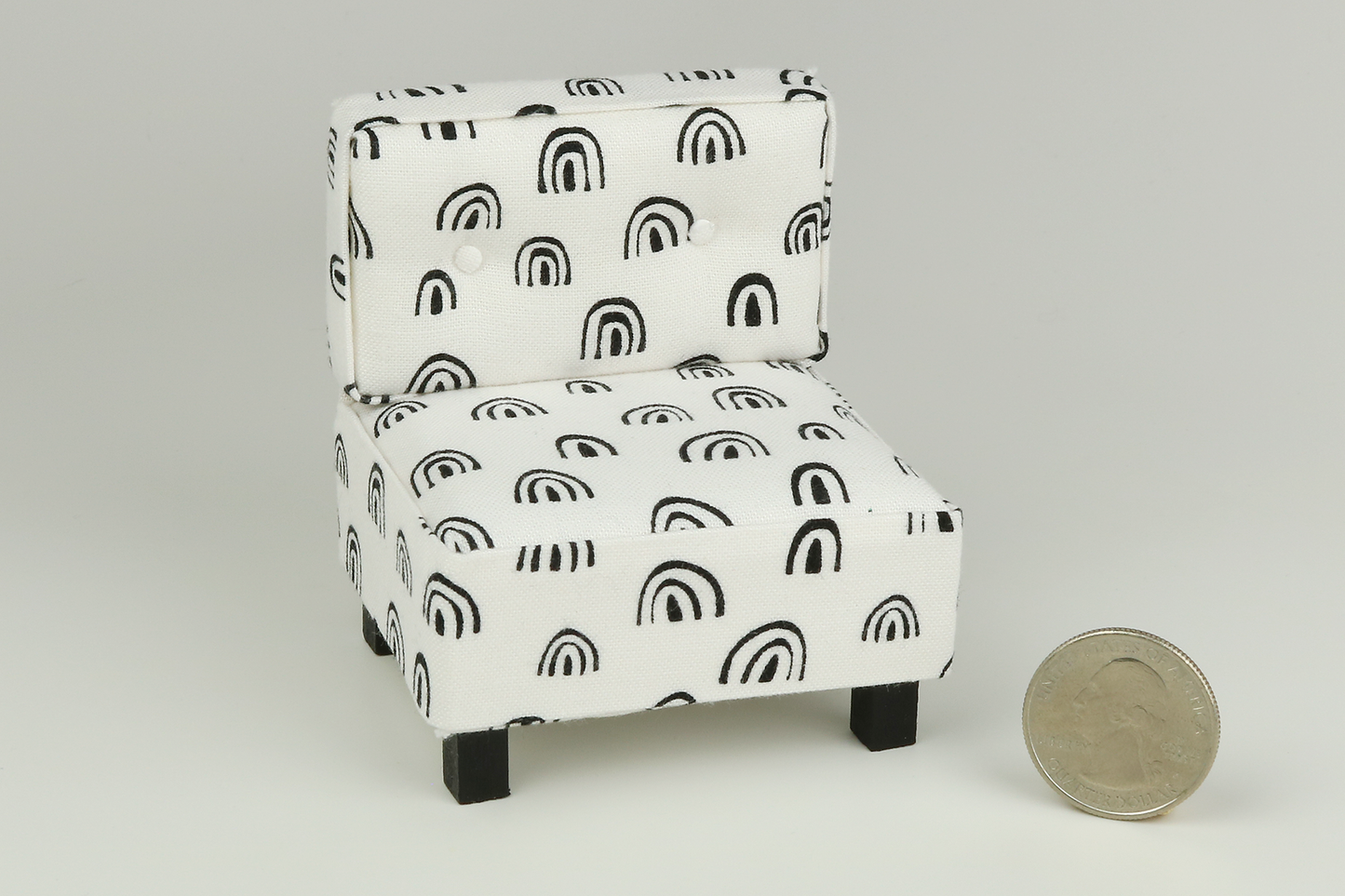 Miniature mid-century modern style side chair with no arms, in a white fabric with black rainbow-shaped patterns throughout, a tufted back with two buttons, and square block walnut legs, next to a quarter for scale on a white background