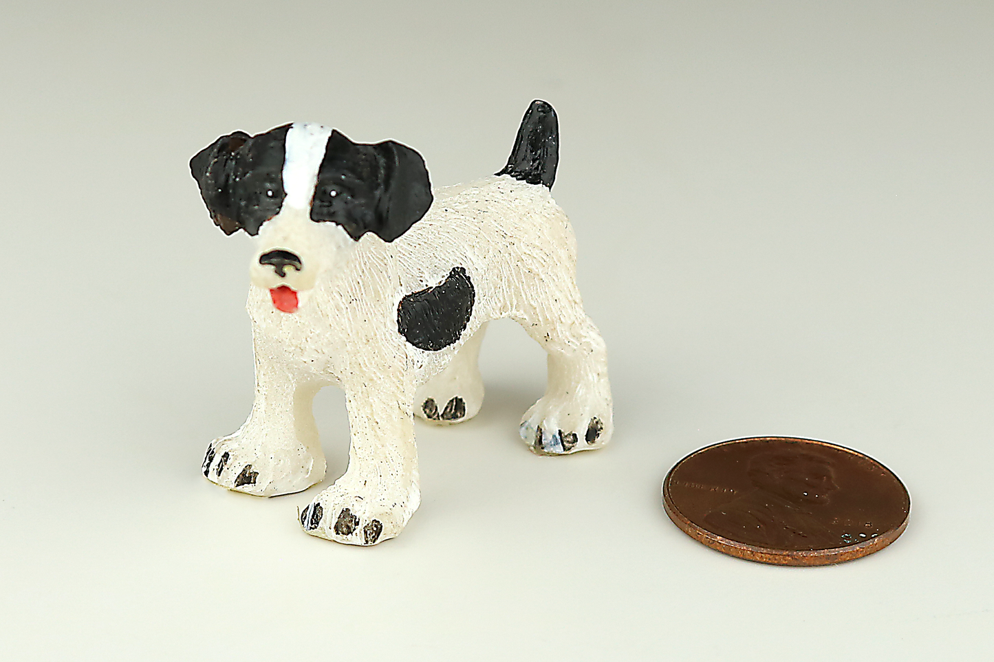 Jack Russell Terrier miniature dog figurine with black and white spots next to a penny on a light gray background