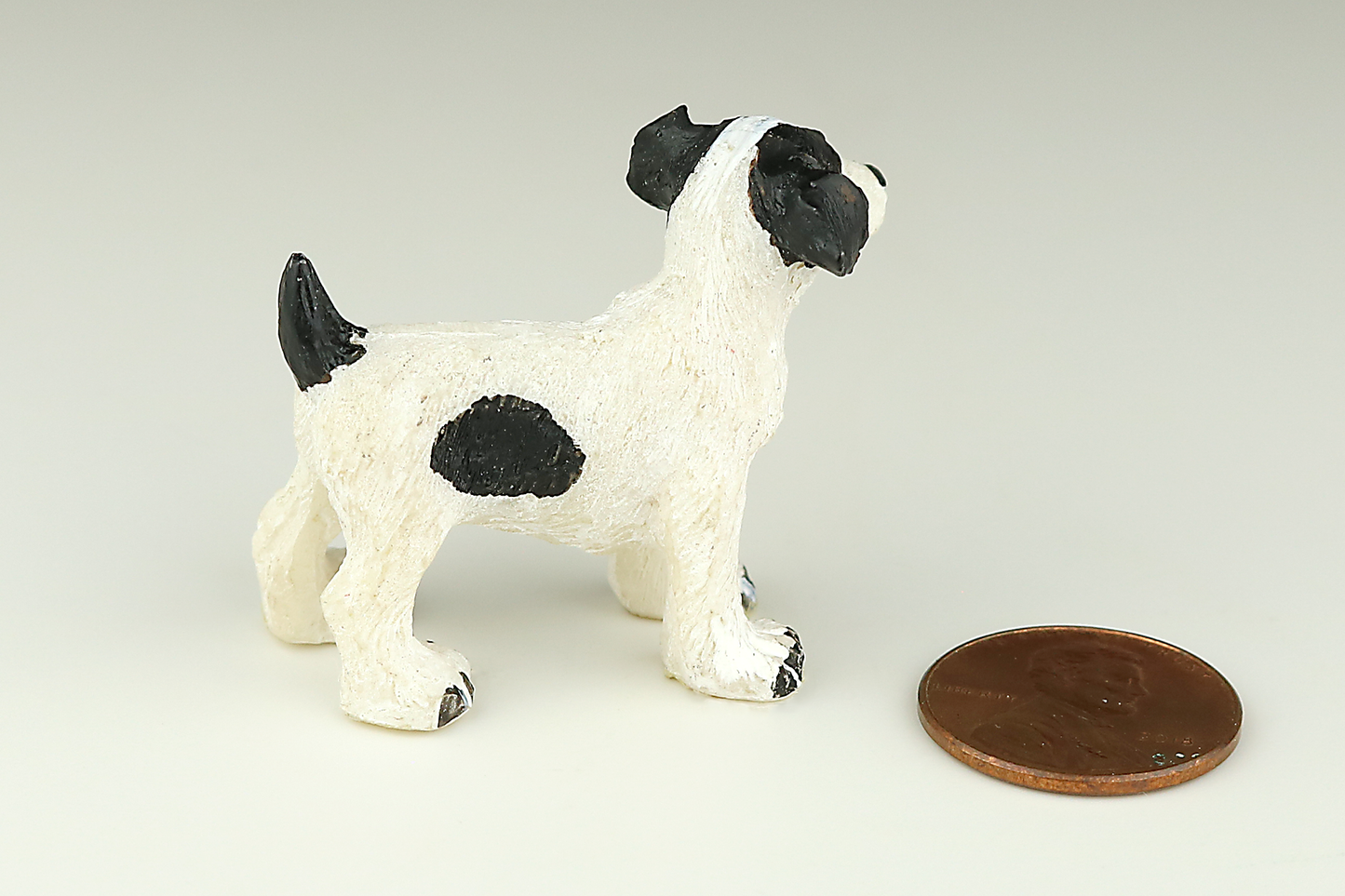 Jack Russell Terrier miniature dog figurine with black and white spots next to a penny on a light gray background