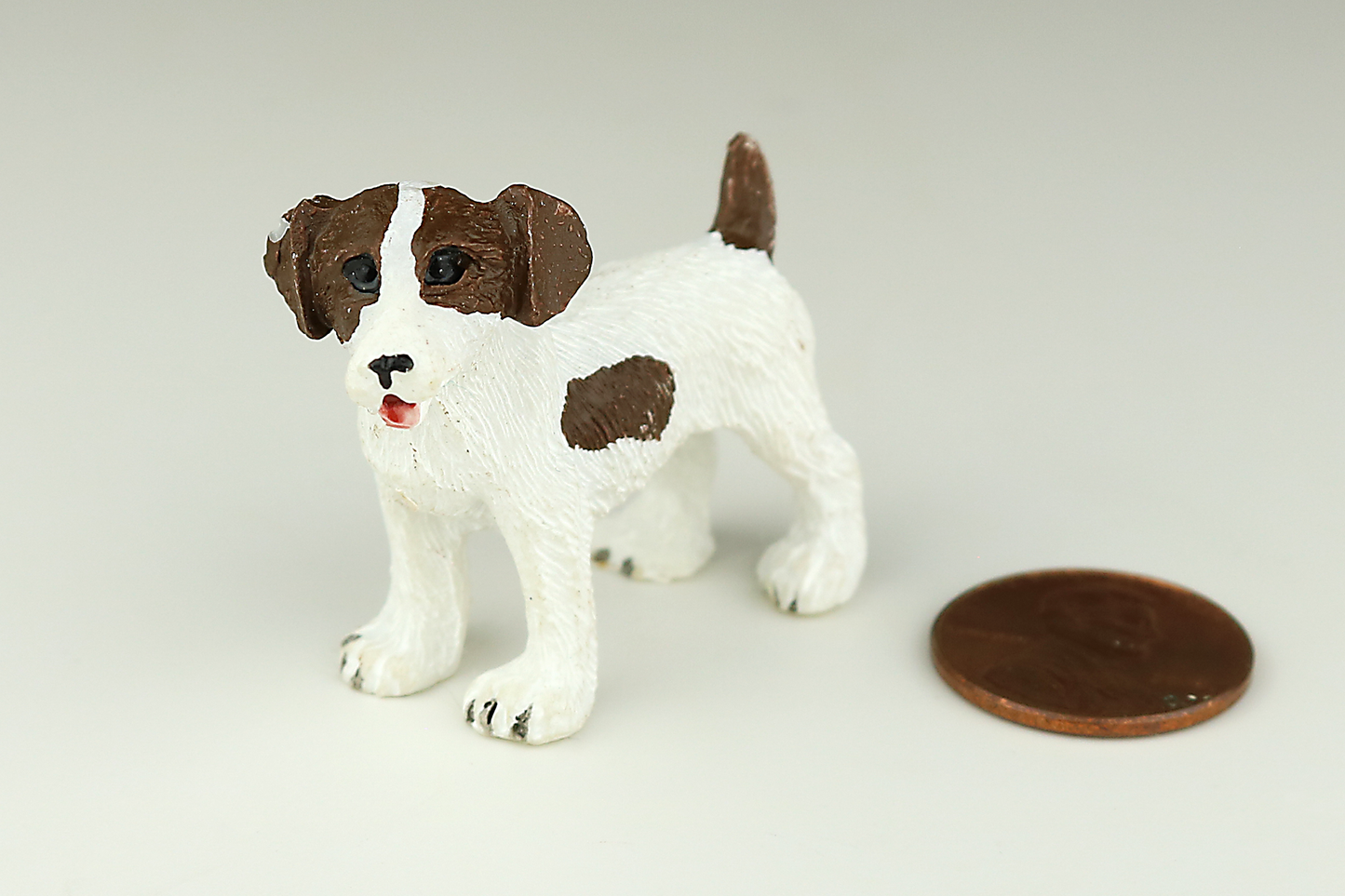 Jack Russell Terrier miniature dog figurine with brown and white spots next to a penny on a light gray background