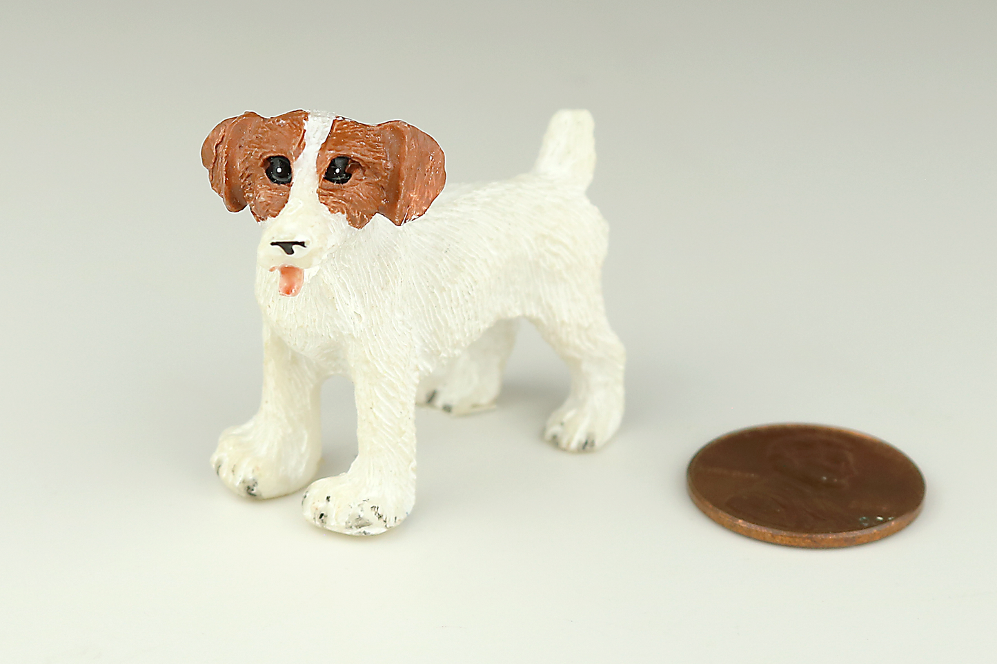 White Jack Russell Terrier miniature dog figurine with red ears next to a penny on a light gray background