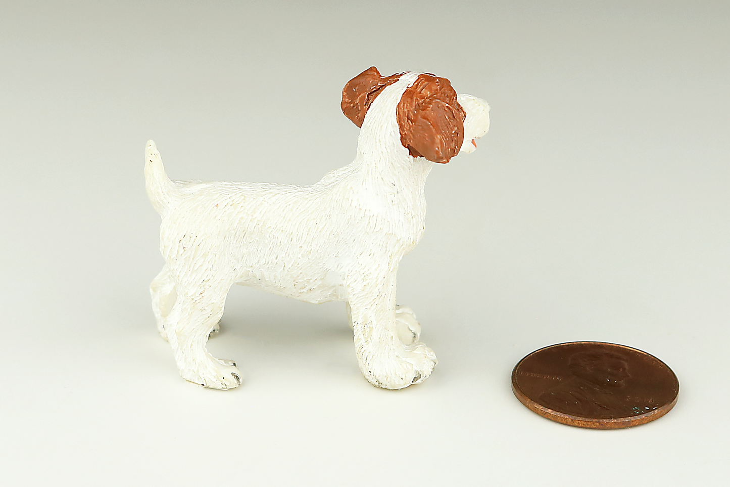 White Jack Russell Terrier miniature dog figurine with red ears next to a penny on a light gray background