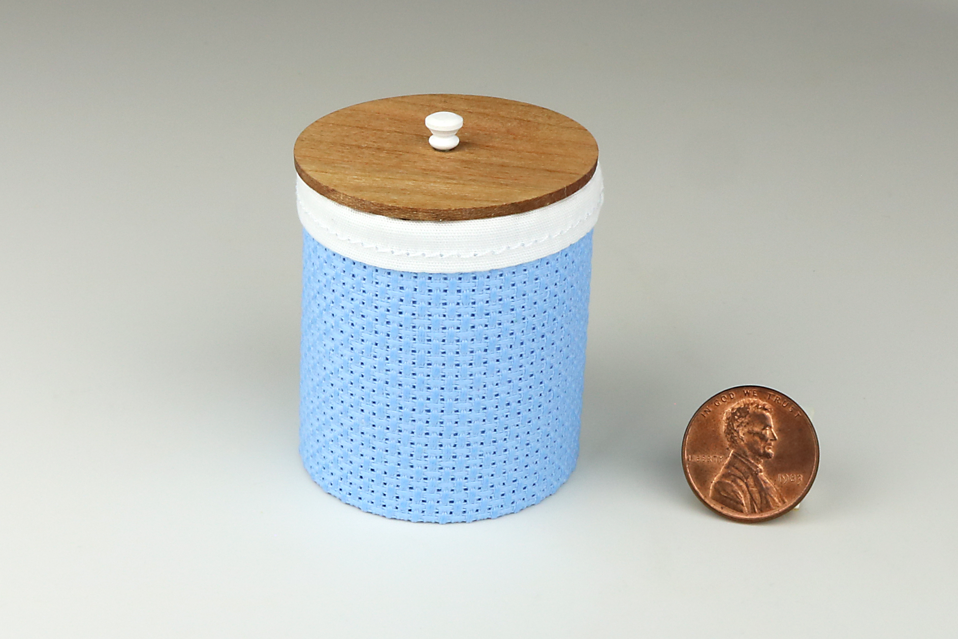 Miniature cylindrical clothes hamper made of a woven material in a medium blue color, with a sewn white cloth insert, a round cherry wood lid, and white handle, pictured next to a penny for scale on a light gray background.