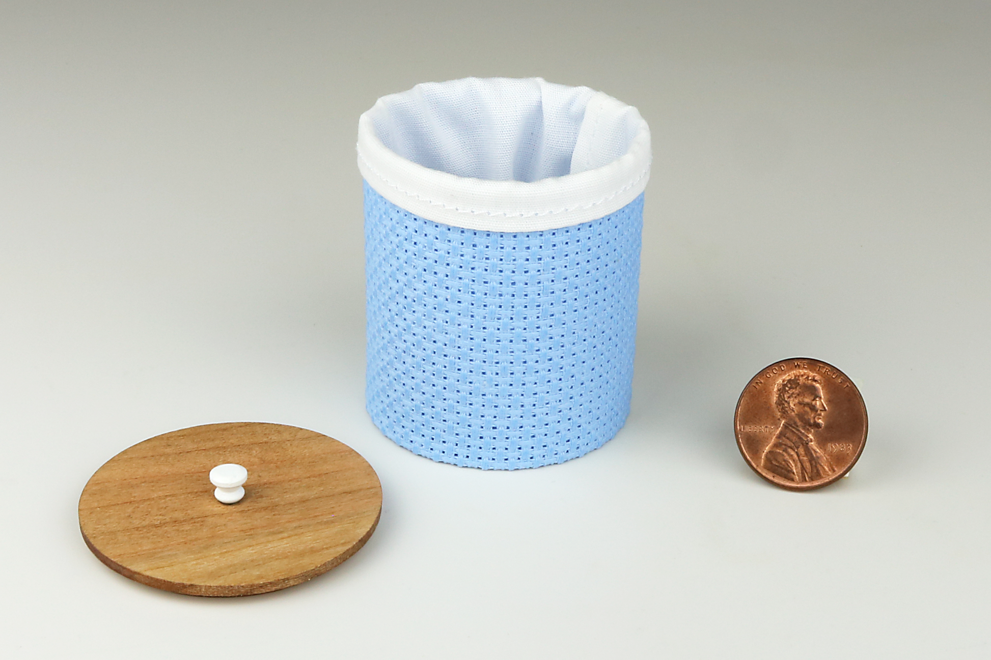 Miniature cylindrical clothes hamper made of a woven material in a medium blue color, open to show the sewn white cloth insert, a round cherry wood lid, and white handle, pictured next to a penny for scale on a light gray background.