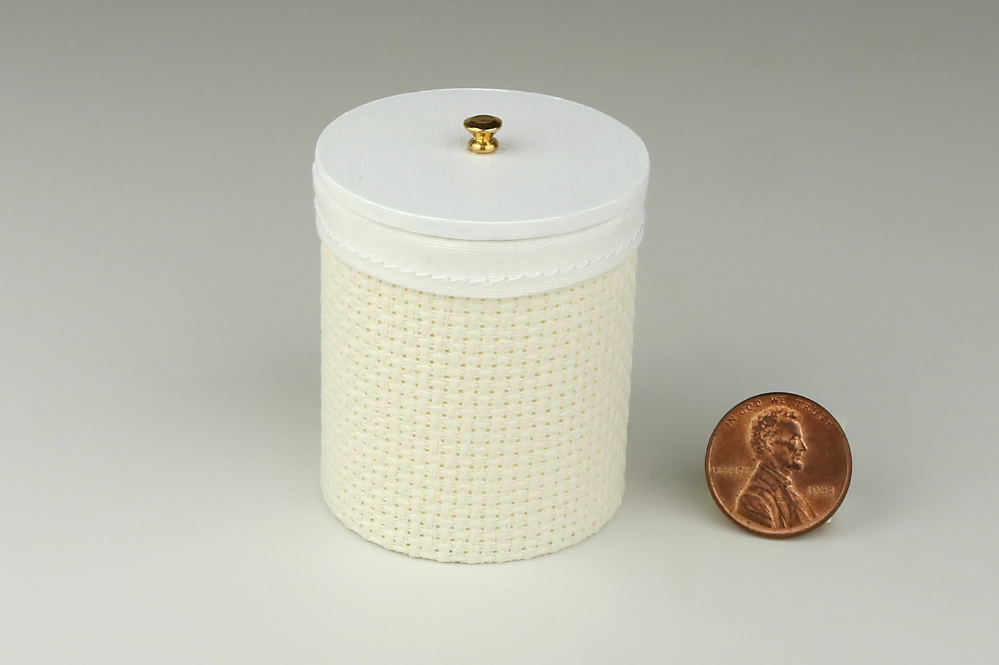 Miniature cylindrical clothes hamper made of a woven material in an ivory color, with sewn white cloth insert, a round white wood lid, and brass handle, pictured next to a penny for scale on a light gray background.