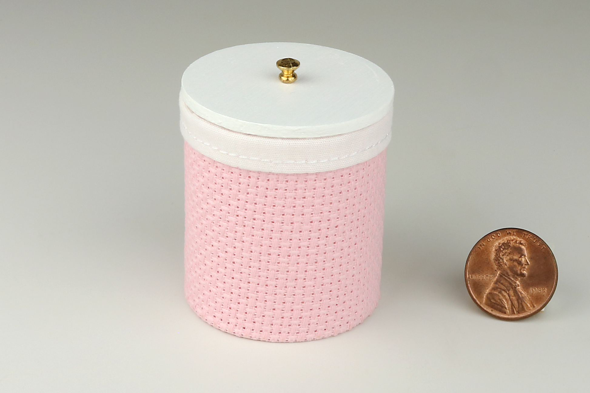Miniature cylindrical clothes hamper made of a woven material in a pale pink color, with a sewn white cloth insert, a round white wood lid, and brass handle, pictured next to a penny for scale on a light gray background.