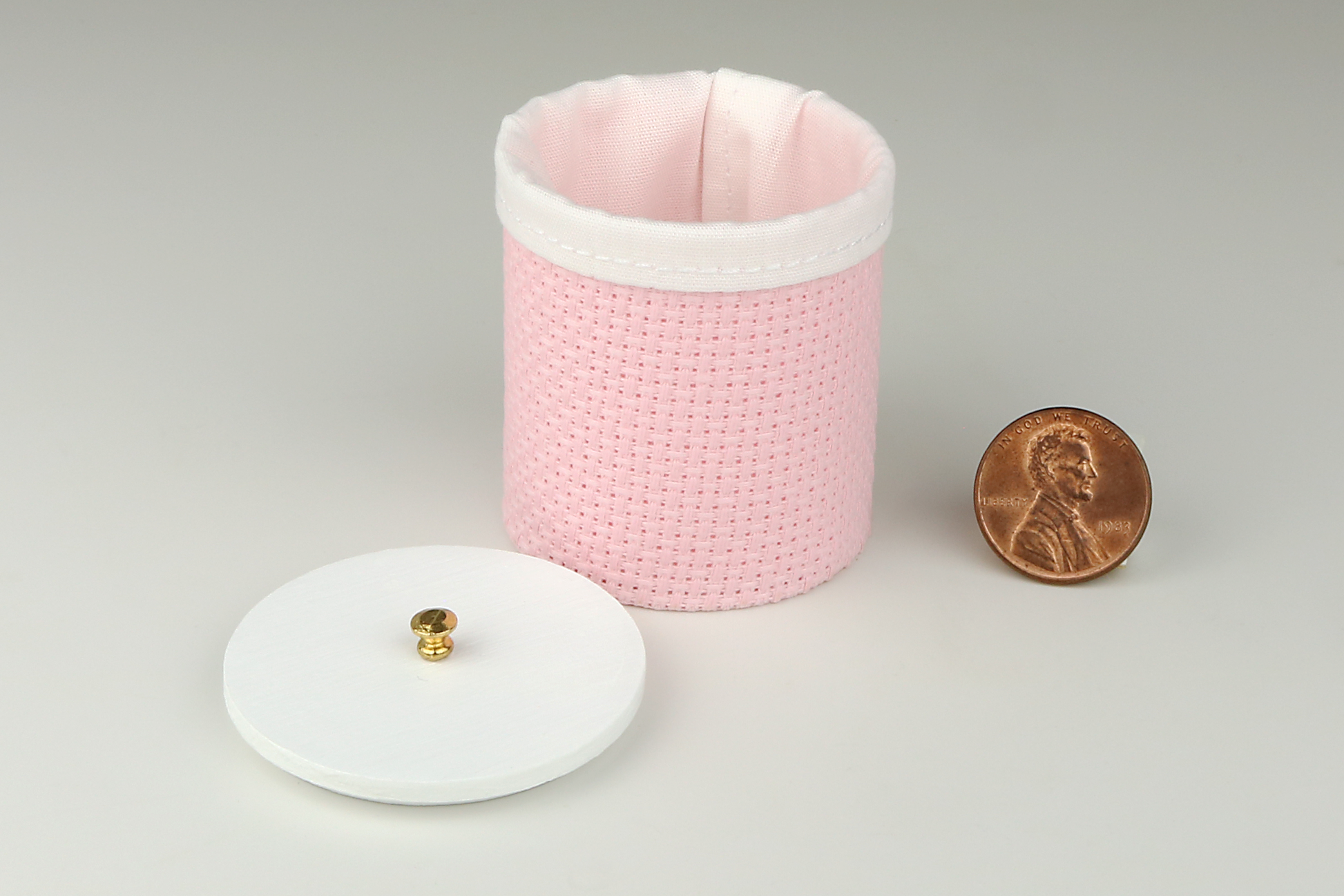 Miniature cylindrical clothes hamper made of a woven material in a pale pink color, open to show the sewn white cloth insert, a round white wood lid, and brass handle, pictured next to a penny for scale on a light gray background.
