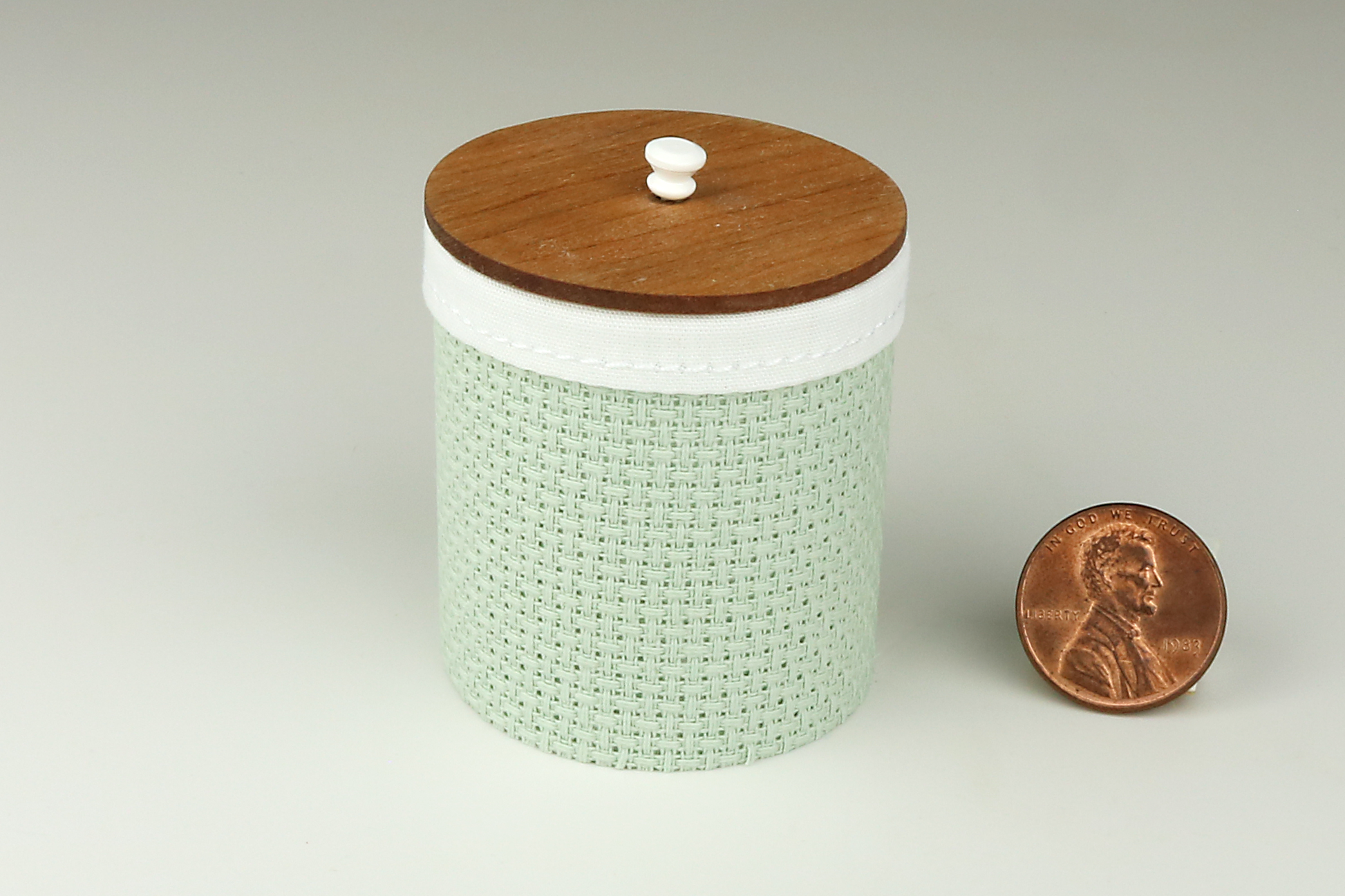 Miniature cylindrical clothes hamper made of a woven material in a sage green color, with a sewn cloth insert, a round cherry wood lid, and white handle, pictured next to a penny for scale on a light gray background.