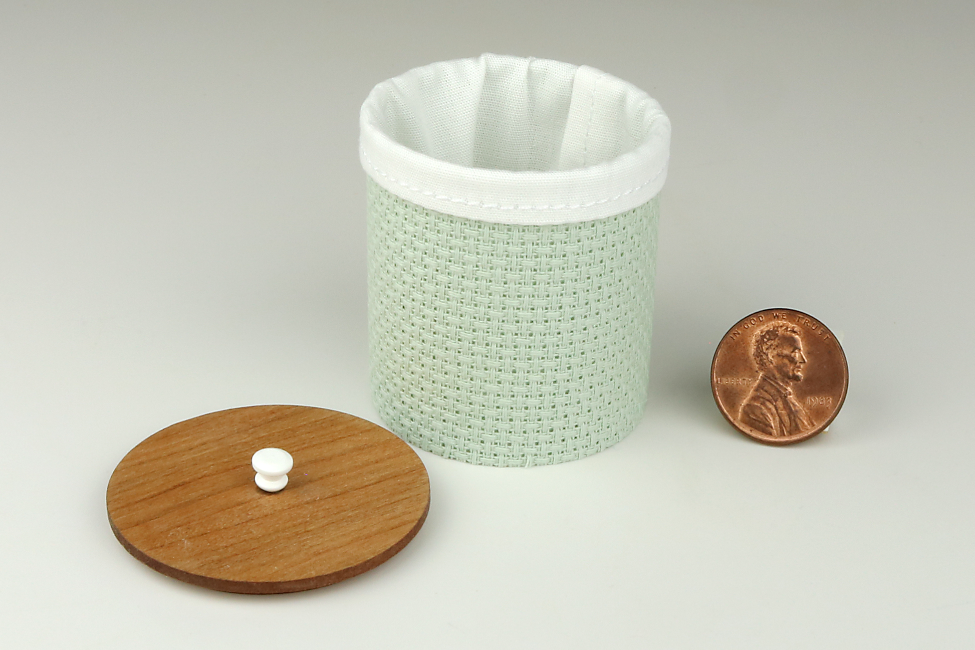 Miniature cylindrical clothes hamper made of a woven material in a sage green color, open to show the sewn white cloth insert, a round cherry wood lid, and white handle, pictured next to a penny for scale on a light gray background.