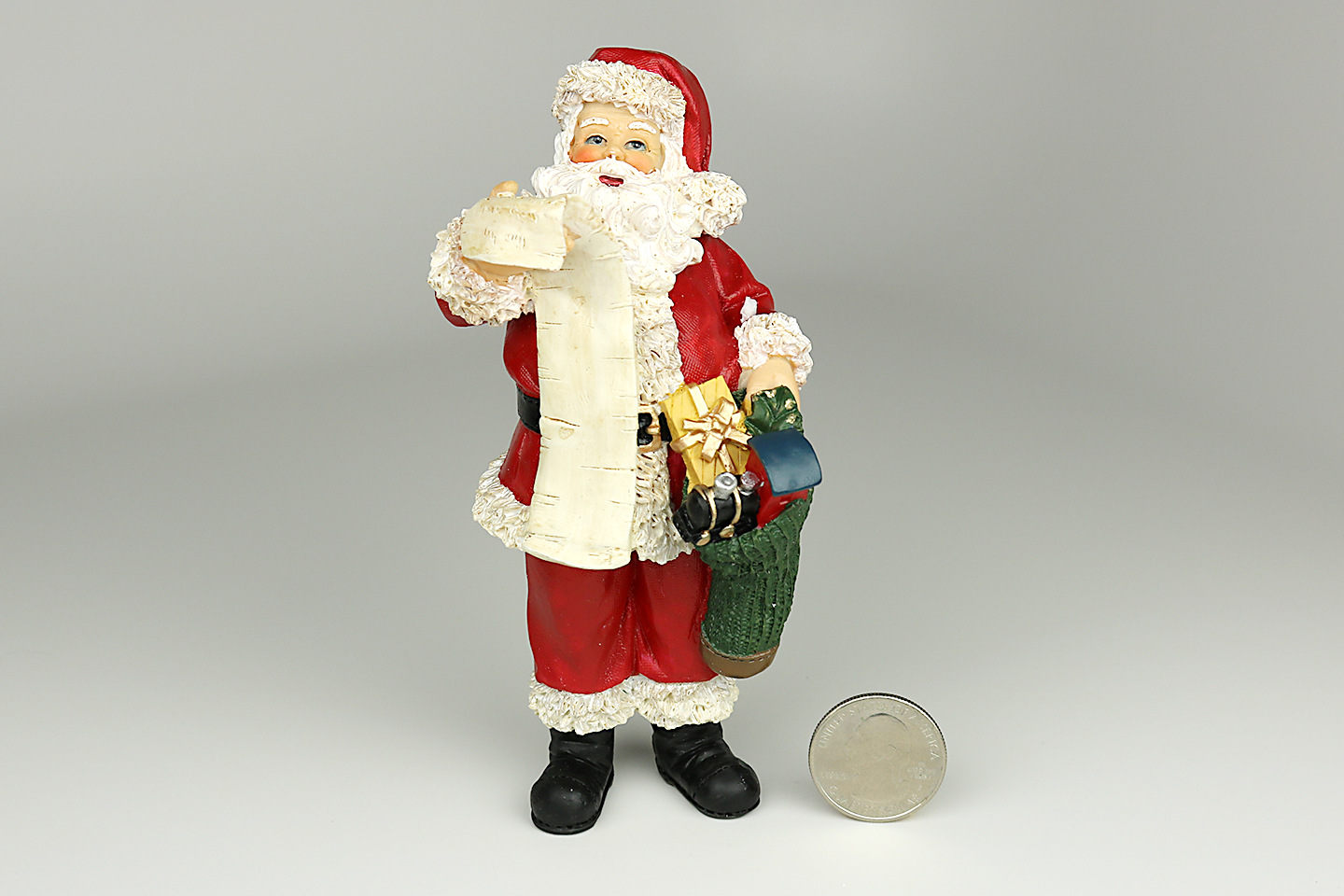 An approximately 6" tall resin figurine of Santa Claus. He is looking up and holding a long, paper list with names on it in his right hand, and a sack of toys in his left hand. An American quarter (25 cent coin) is next to his feet to demonstrate scale.