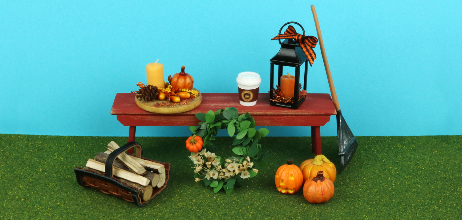 A miniature red rustic park bench with several autumn-themed miniature products surrounding it, on a blue background with green grass at the bottom.