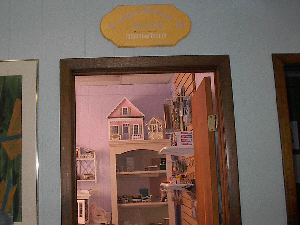 A dated-looking photo of a doorway looking into a room painted purple, with several dollhouse miniatures organized neatly inside on shelves, a pegboard and a bookcase, on top of which is a pink chalet-style dollhouse. Above the doorway is a yellow plaque with "Dollhouse Alley Miniature Houses" hand painted in purple, and a stenciled-on white picket fence.