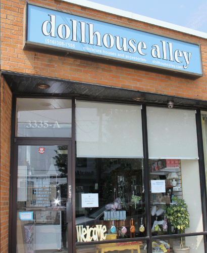 Brick storefront of Dollhouse Alley during the day, with a blue sign bearing the business name and information. There are several cute items for sale that are semi-visible inside the store through the glass windows and door, and a small decorative welcome sign propped in the window.