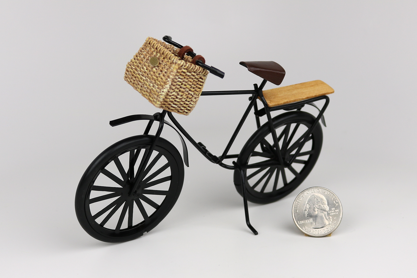 Miniature black bicycle with a basket next to a quarter for scale on a light gray background.