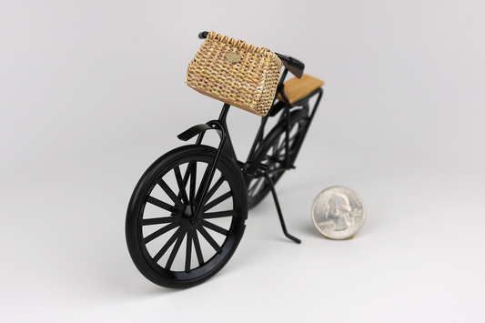Miniature black bicycle with a basket next to a quarter for scale on a light gray background.