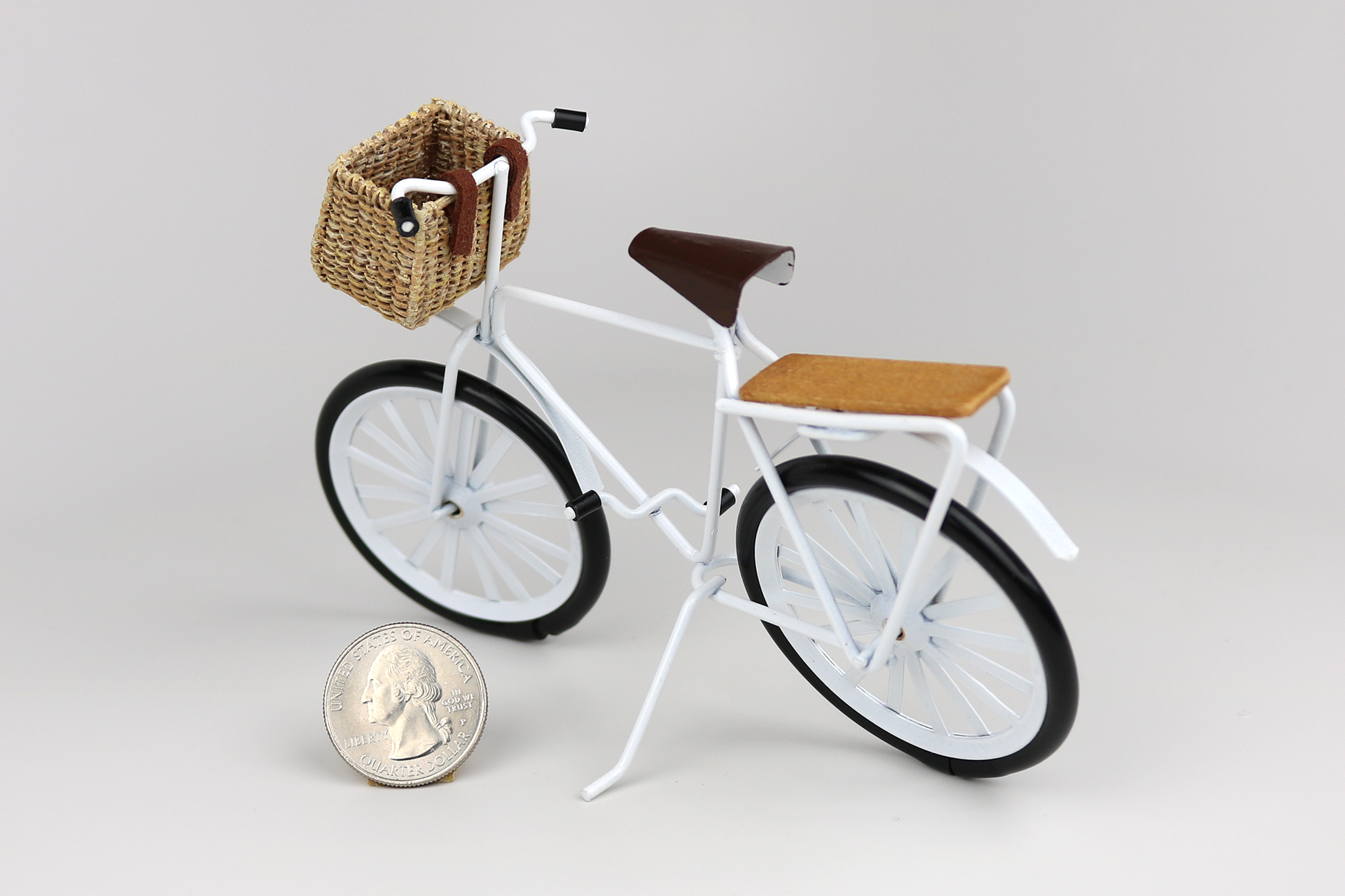 Miniature white bicycle with a basket next to a quarter for scale on a light gray background.