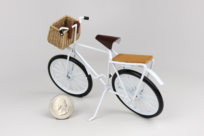 Miniature white bicycle with a basket next to a quarter for scale on a light gray background.