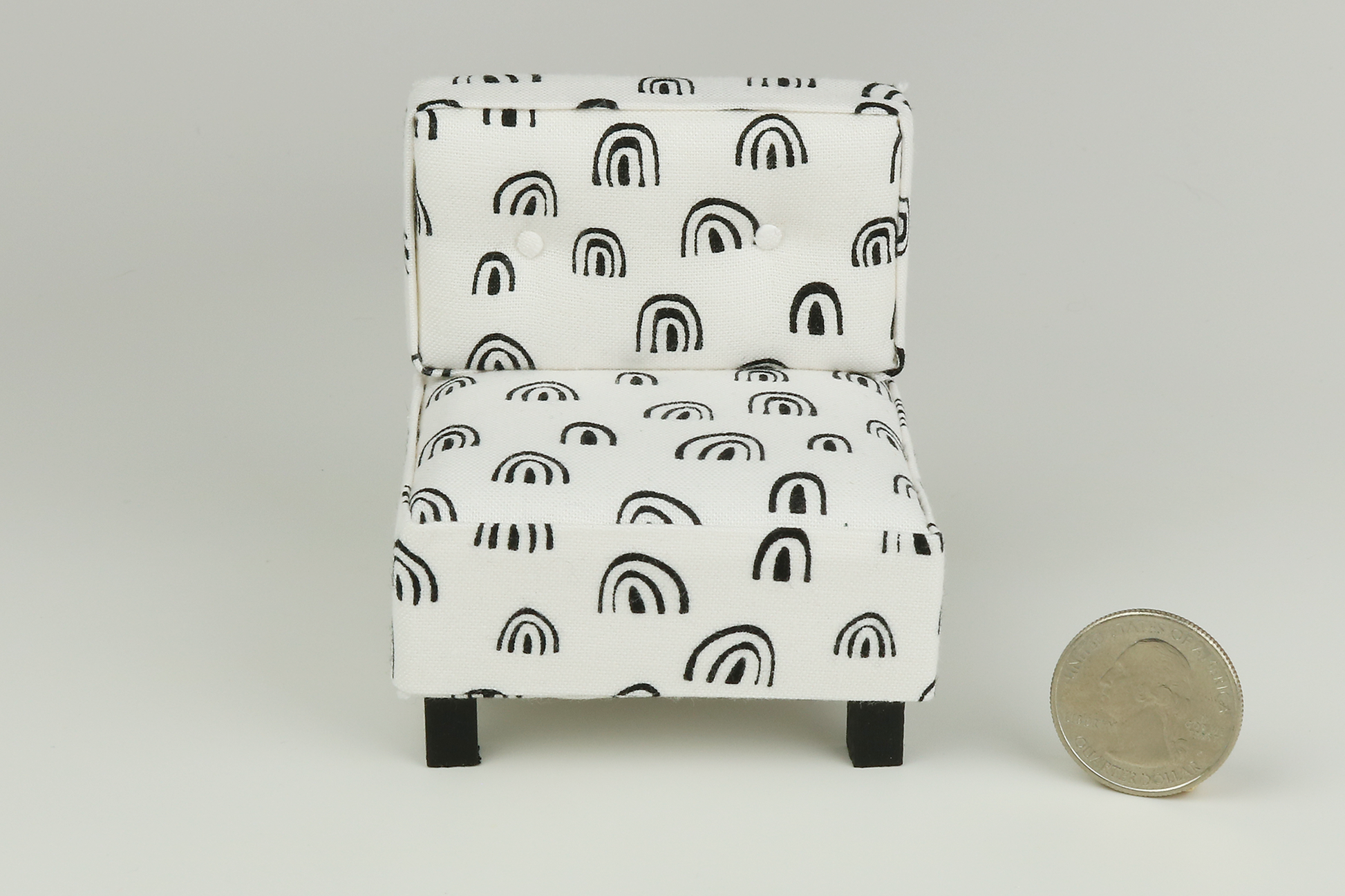 Miniature mid-century modern style side chair with no arms, in a white fabric with black rainbow-shaped patterns throughout, a tufted back with two buttons, and square block walnut legs, next to a quarter for scale on a white background