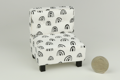 Miniature mid-century modern style side chair with no arms, in a white fabric with black rainbow-shaped patterns throughout, a tufted back with two buttons, and square block walnut legs, next to a quarter for scale on a white background