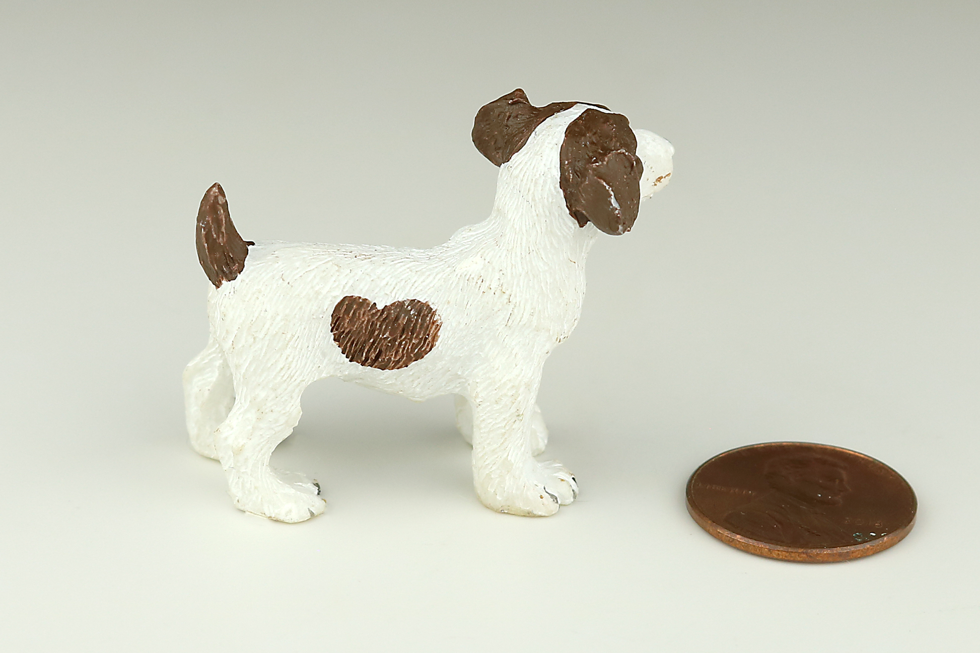Jack Russell Terrier miniature dog figurine with brown and white spots next to a penny on a light gray background