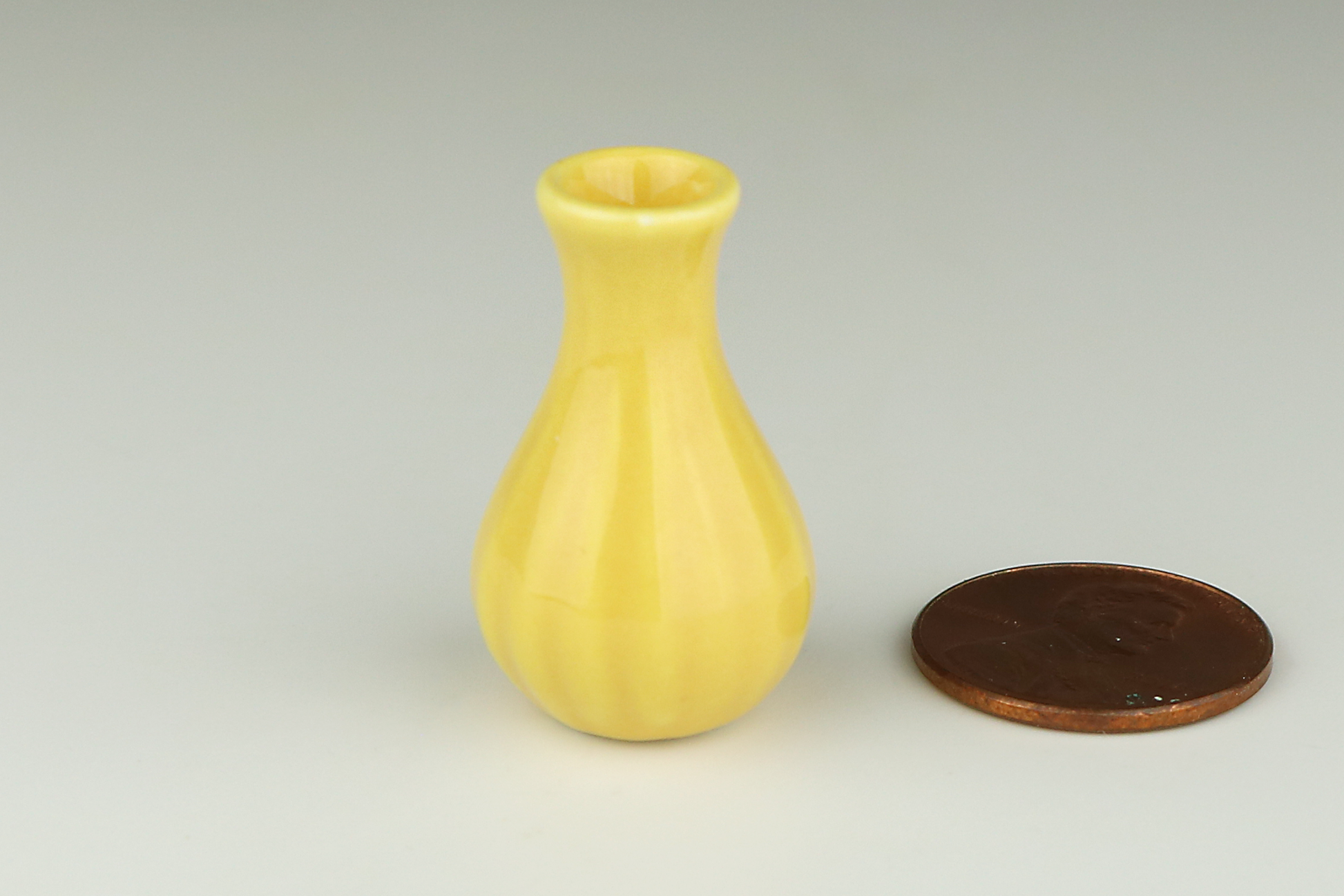 Miniature yellow ceramic vase, wider on the bottom, tapered in at the top, with a slightly fluted texture, next to a penny on a white background
