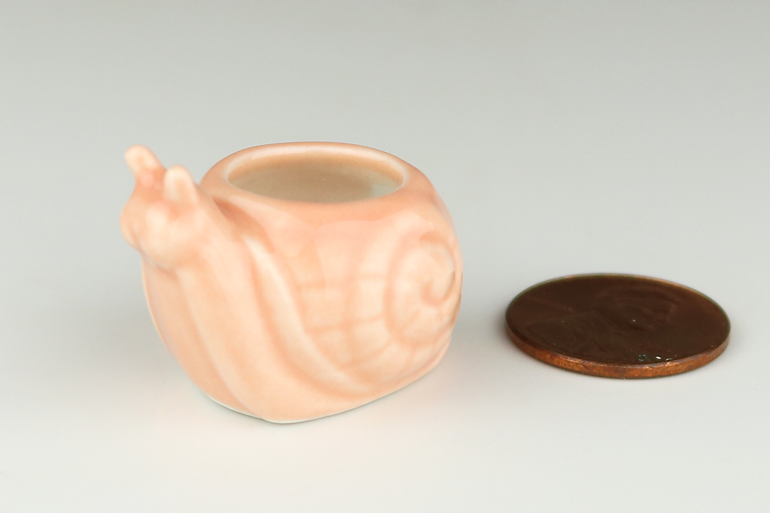 Miniature peach-colored ceramic snail-shaped planter on next to a penny on a light gray background