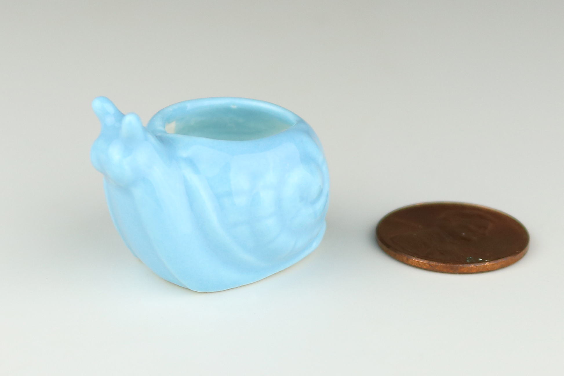 Miniature pale-blue ceramic snail-shaped planter on next to a penny on a light gray background