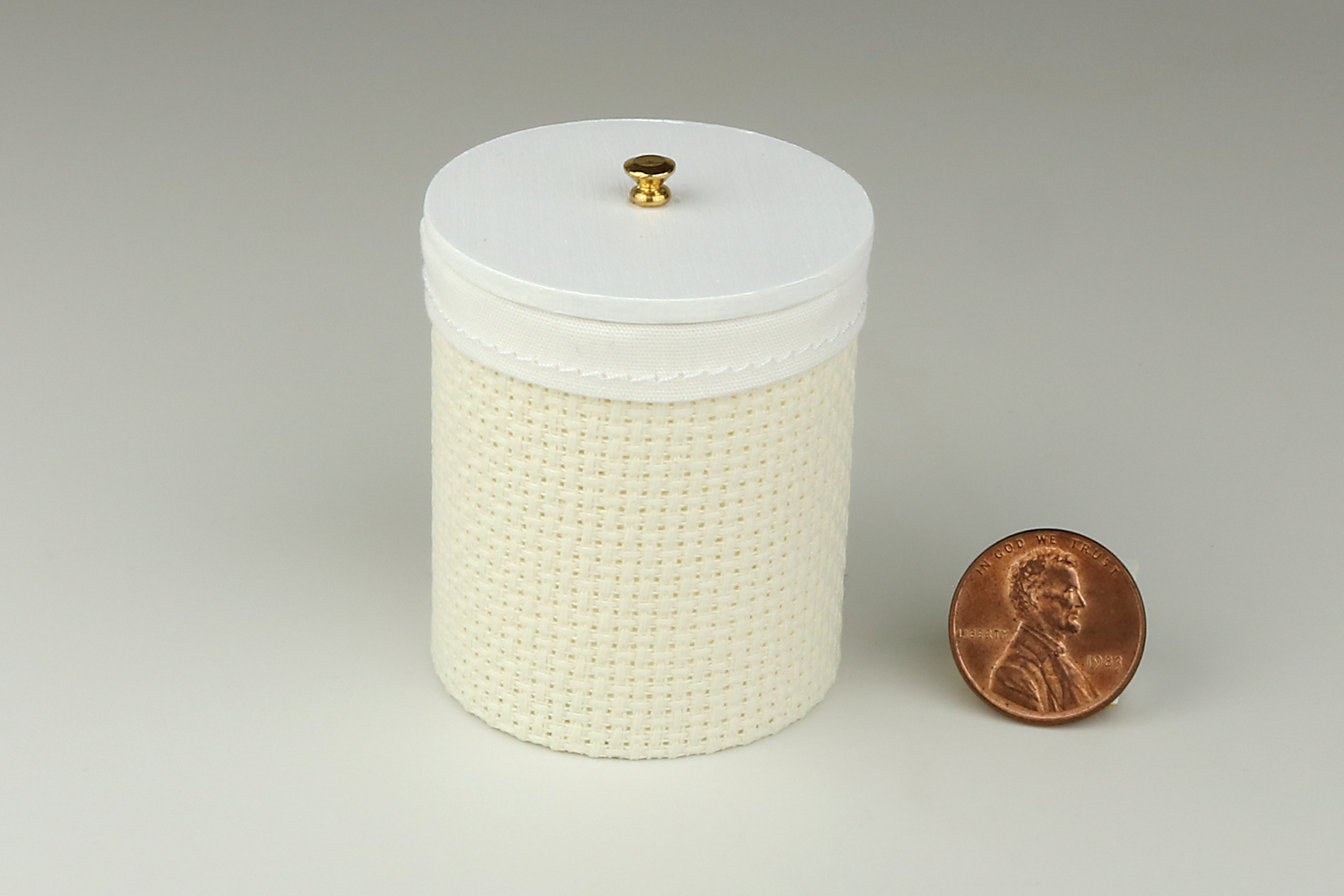 Miniature cylindrical clothes hamper made of a woven material in an ivory color, with sewn white cloth insert, a round white wood lid, and brass handle, pictured next to a penny for scale on a light gray background.