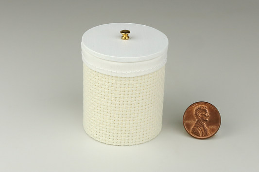Miniature cylindrical clothes hamper made of a woven material in an ivory color, with sewn white cloth insert, a round white wood lid, and brass handle, pictured next to a penny for scale on a light gray background.