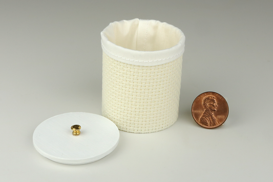 Miniature cylindrical clothes hamper made of a woven material in an ivory color, open to show the sewn white cloth insert, a round white wood lid, and brass handle, pictured next to a penny for scale on a light gray background.
