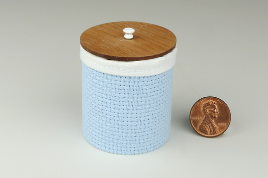 Miniature cylindrical clothes hamper made of a woven material in a sage green color, with a sewn white cloth insert, a round cherry wood lid, and white handle, pictured next to a penny for scale on a light gray background.