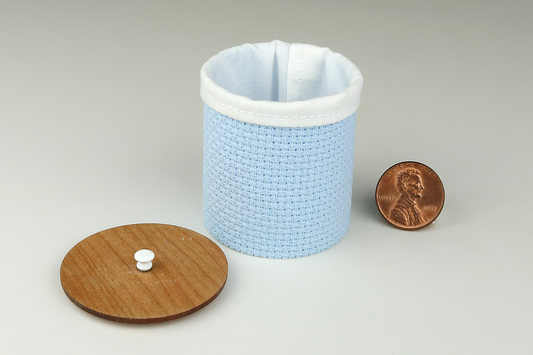 Miniature cylindrical clothes hamper made of a woven material in a light blue color, open to show the sewn white cloth insert, a round cherry wood lid, and white handle, pictured next to a penny for scale on a light gray background.