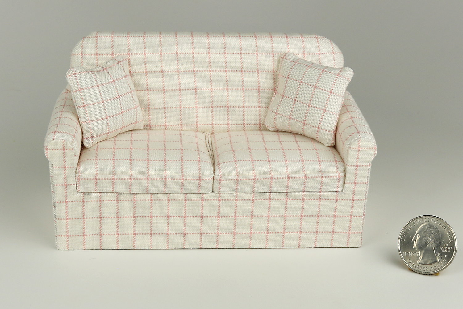 Miniature sofa in a pale pink plaid print on a cream background, with matching pillows, pictured on a gray background, next to a quarter for scale.
